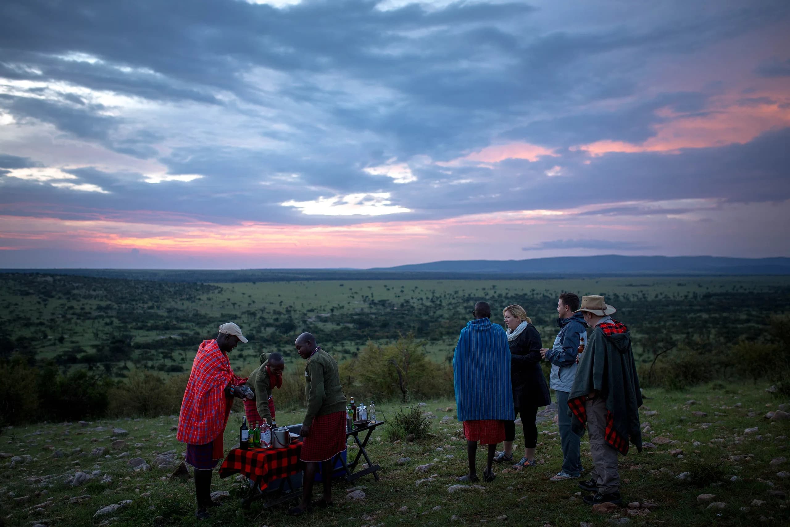 A safari guide leaning on an open 4x4 door, talking with two travellers at the edge of a savannah track